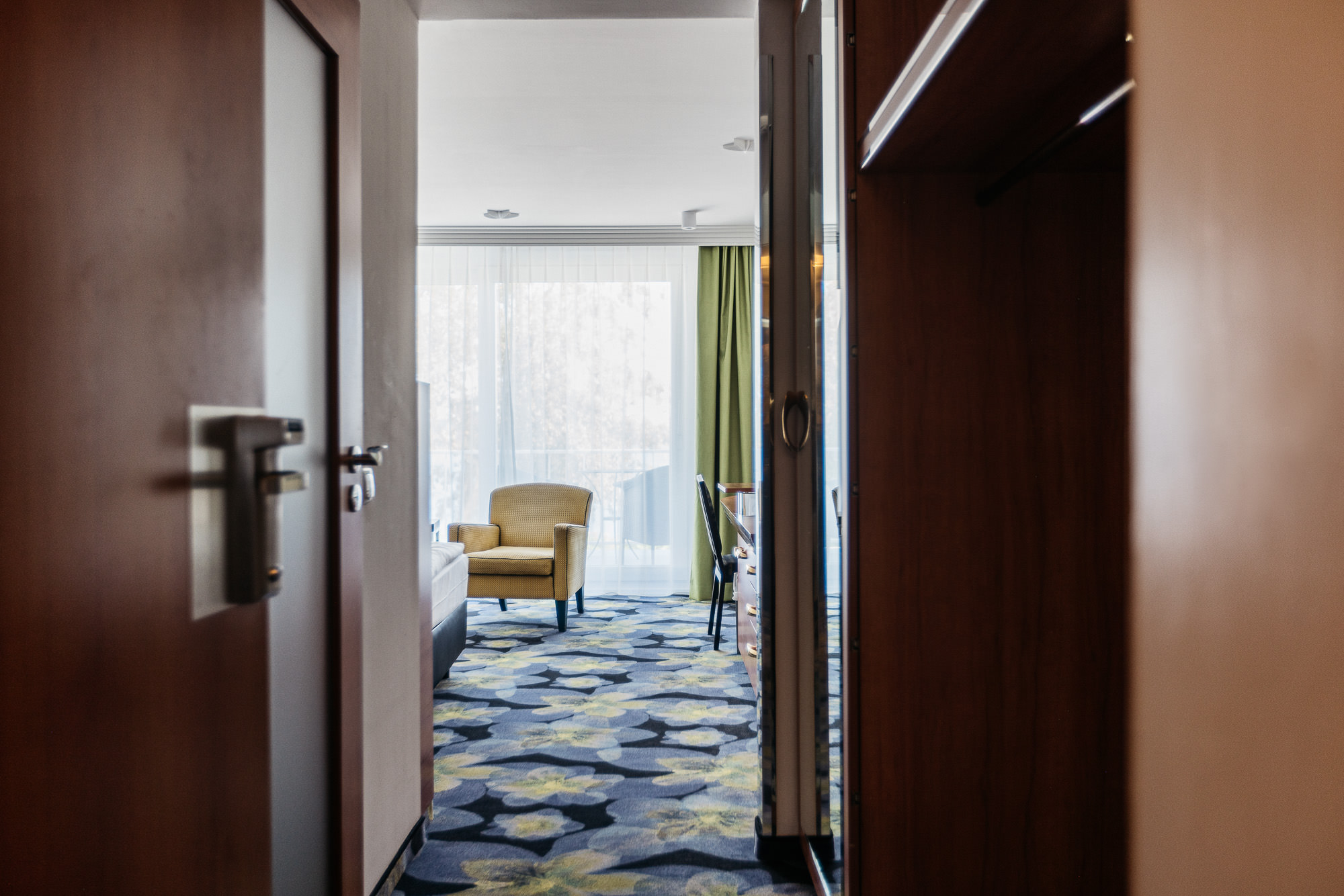 Eine offene Hotelzimmertür gibt Einblick in den Flur und den hinteren Bereichs eines Doppelzimmers mit einem Sessel vor der Fensterfront und geschlossenen weißen Vorhängen.