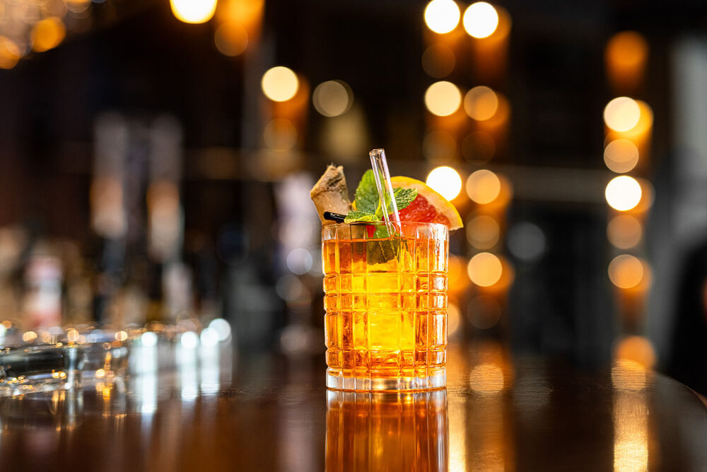 Orange leuchtender Cocktail steht in einem edlem Whiskeyglas mit Strohhalm und Zitrone stehen auf dem Tresen mit der unscharfen Leonardo Bar im Hintergrund. 