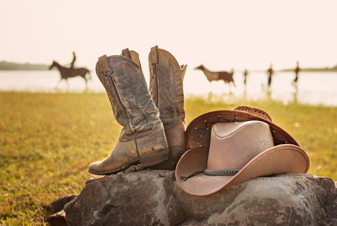 Typische Cowboy Stiefel und Cowboy Hut liegen szenisch auf einer Wiese vor einem See.