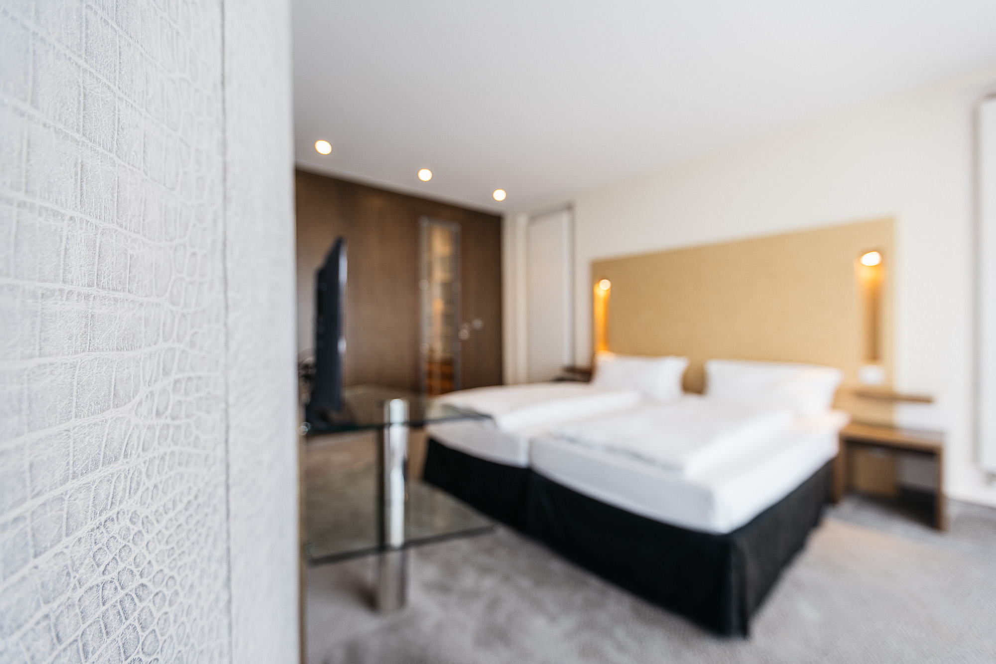 Ein großes Doppel-Boxspringbett steht an einer Wand neben einer großen Fensterfront und einer Tapete in heller Lederoptik.